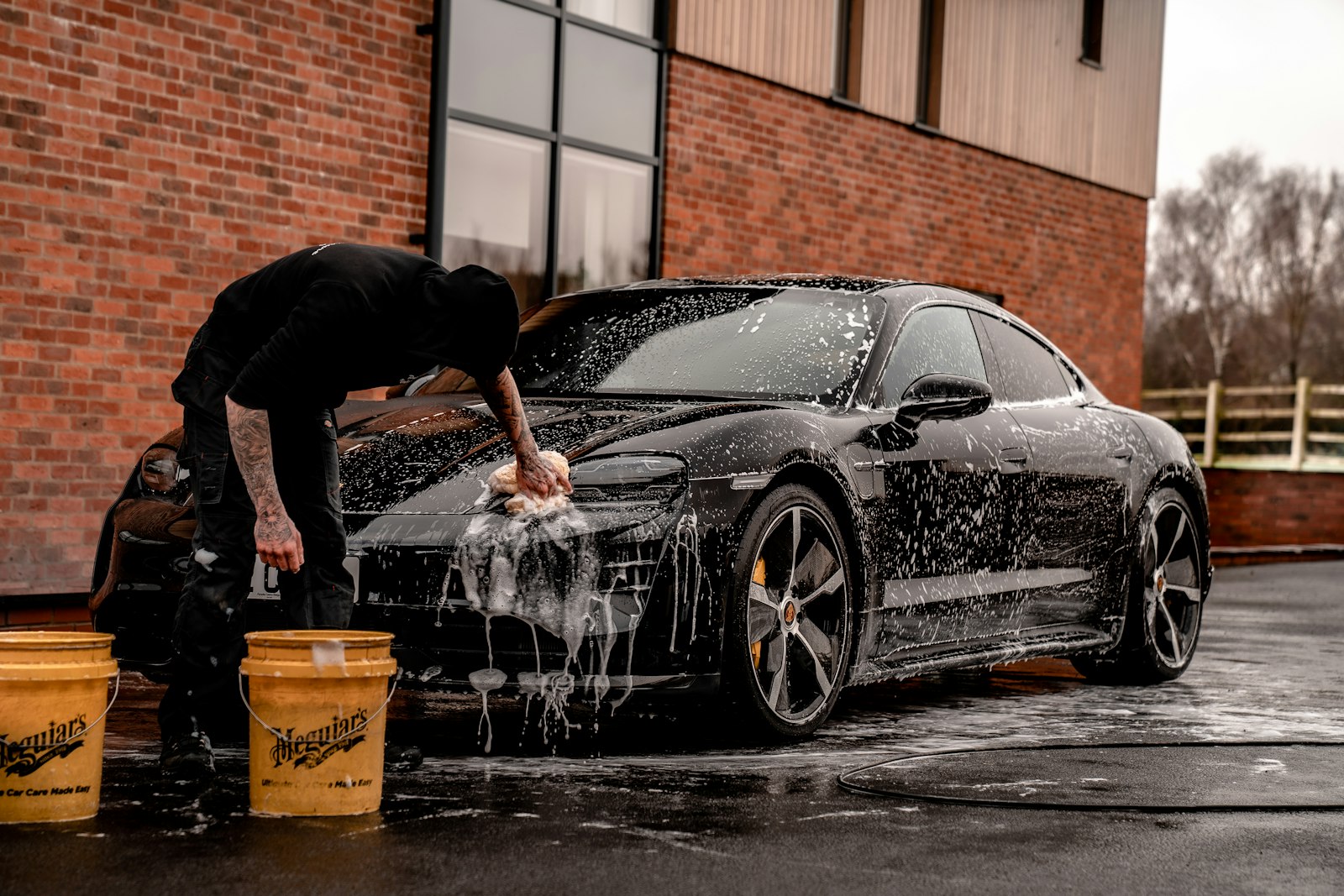 A vehicle being hand-washed with foam and two buckets outside a brick building.